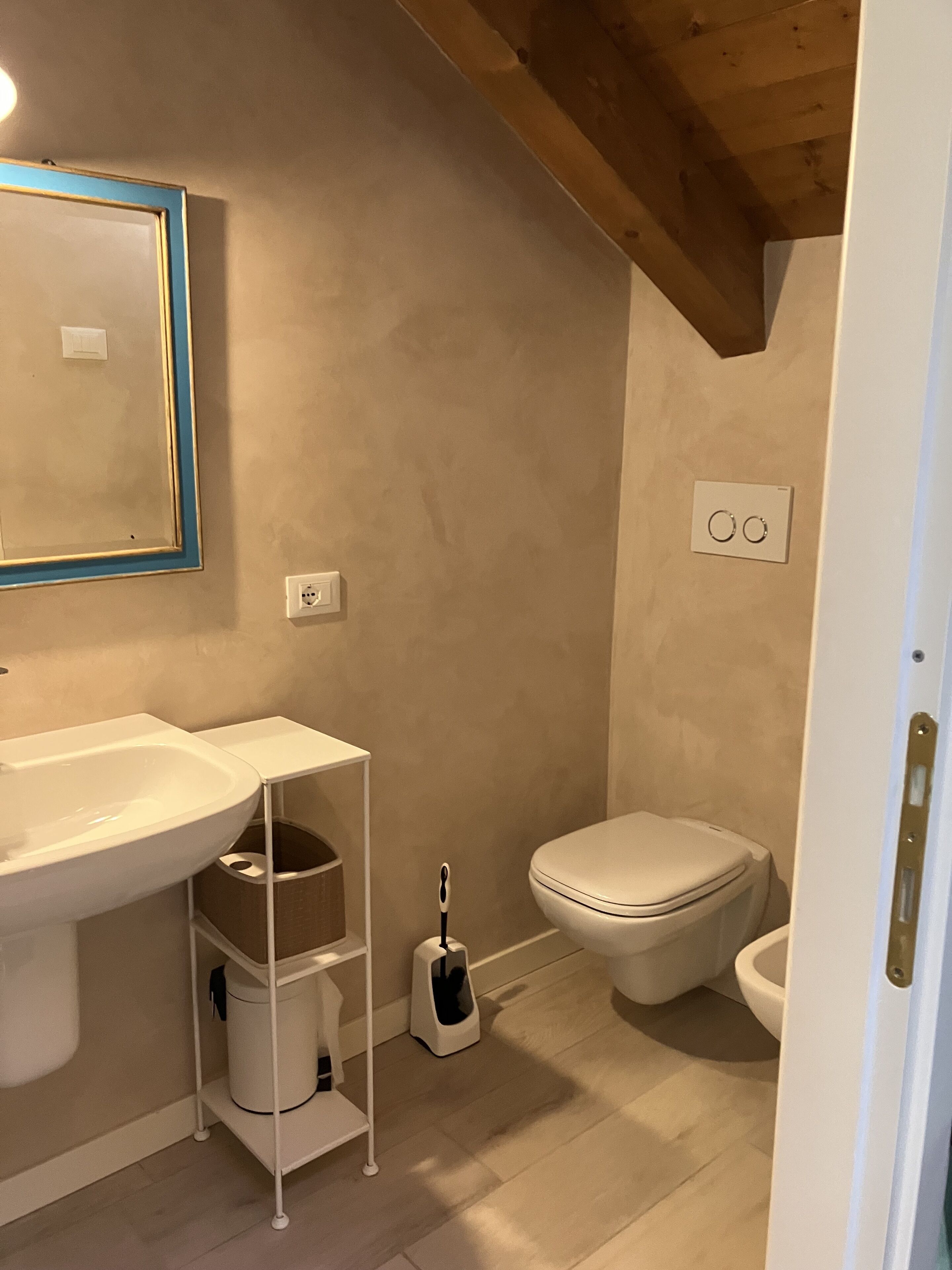 A bathroom with a white sink, mirror, and toilet. There is a small shelf with a trash bin and a toilet brush.