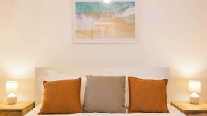 Een hotelkamer met een bed, drie kussens, een nachtkastje met een lamp en een ingelijst foto aan de muur.