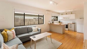 Een modern appartement met een grijze bank, een witte salontafel, een keuken met witte kastjes en een groot raam met zonwering.