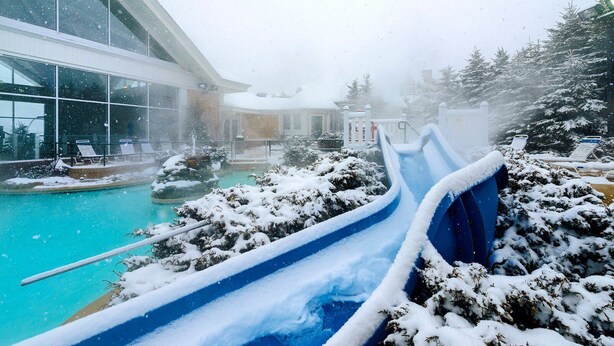 Un espace extérieur enneigé comprenant une piscine, un toboggan bleu et un bâtiment doté de grandes fenêtres.