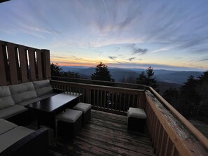 Um terraço com sofá, mesa e cadeiras, com vista para uma paisagem montanhosa ao pôr do sol.