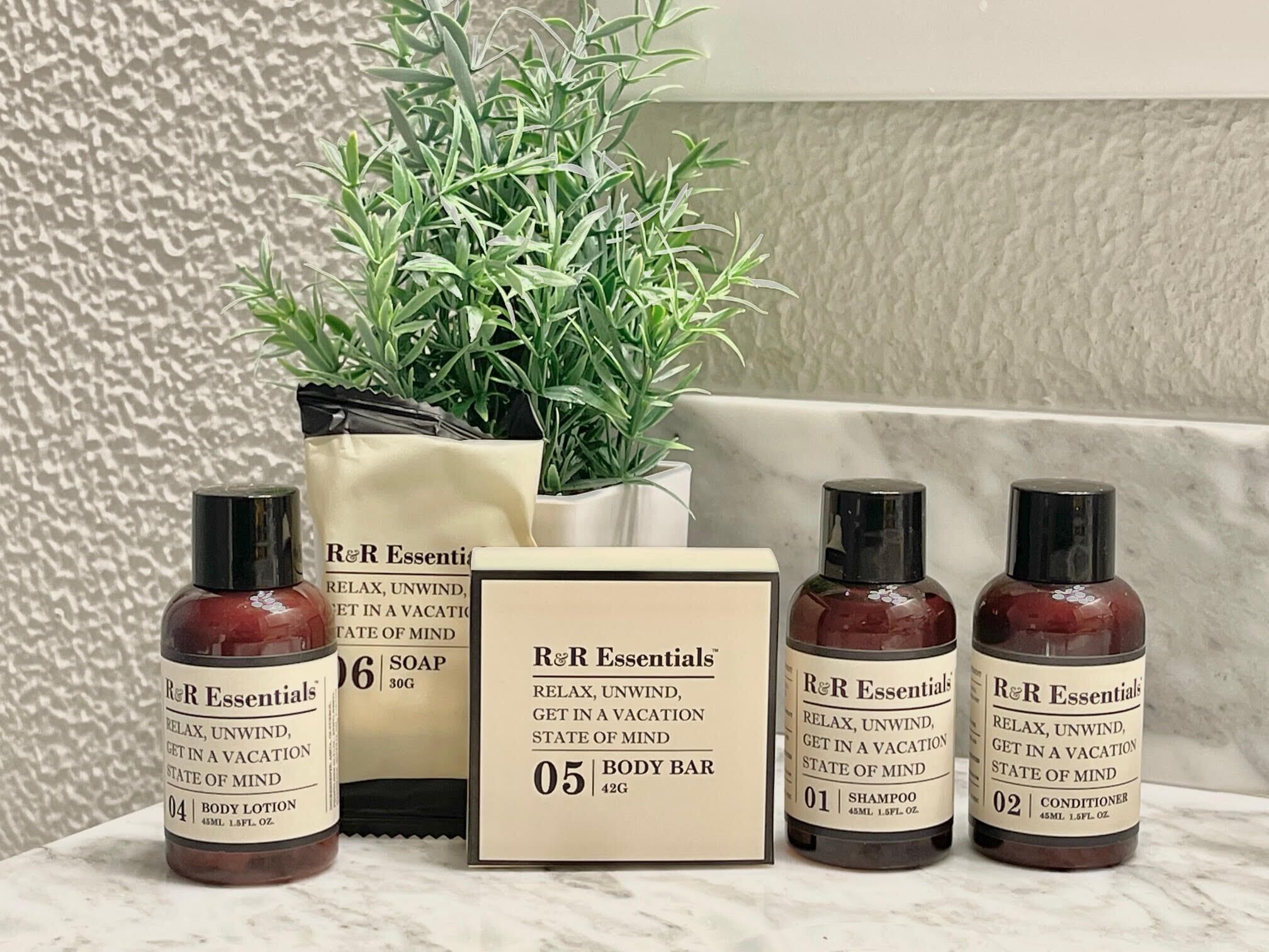 A bathroom with a marble countertop, a potted plant, and four bottles of R&R Essentials body products.