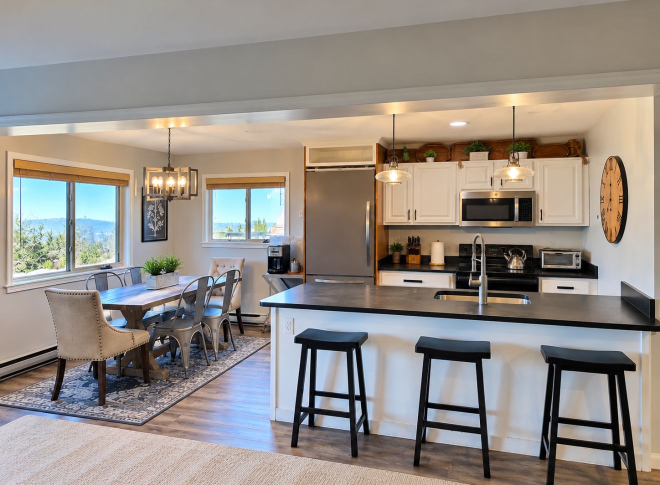 A modern kitchen with a central island, bar stools, a dining area with a table and chairs, and a view of the outdoors.
