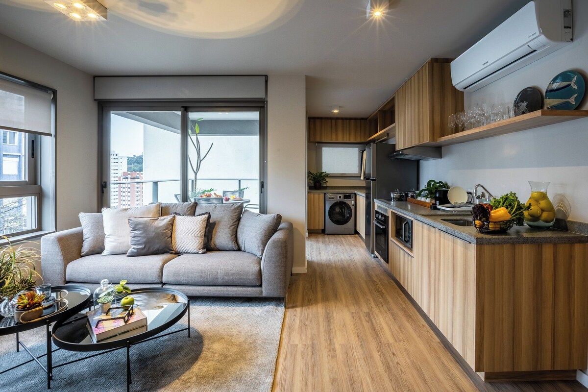 Apartamento moderno com sala de estar, sofá cinza, mesa de centro e vista para uma varanda por meio de uma porta de vidro deslizante. A cozinha possui armários de madeira, forno embutido e pia.