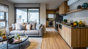 Un apartamento moderno con una sala de estar que incluye un sofá gris, una mesita de centro y vistas a un balcón a través de un ventanal corredero. La cocina tiene armarios de madera, un horno empotrado y un lavamanos.