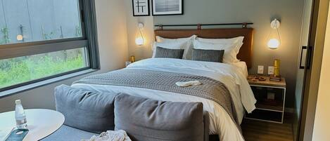 A hotel room with a bed, a small table with slippers and a water bottle, a bedside table with a lamp, and framed pictures on the wall.