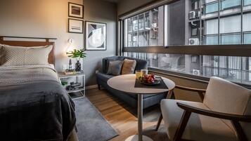 Een moderne hotelkamer met een bed, een bank, een ronde tafel met fruit en een groot raam met uitzicht op de stad.