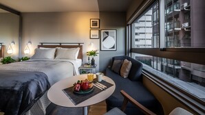Una habitación de hotel moderna con una cama grande, un sofá, una mesa de comedor con frutas y una bebida, y vistas de la ciudad a través de amplios ventanales.
