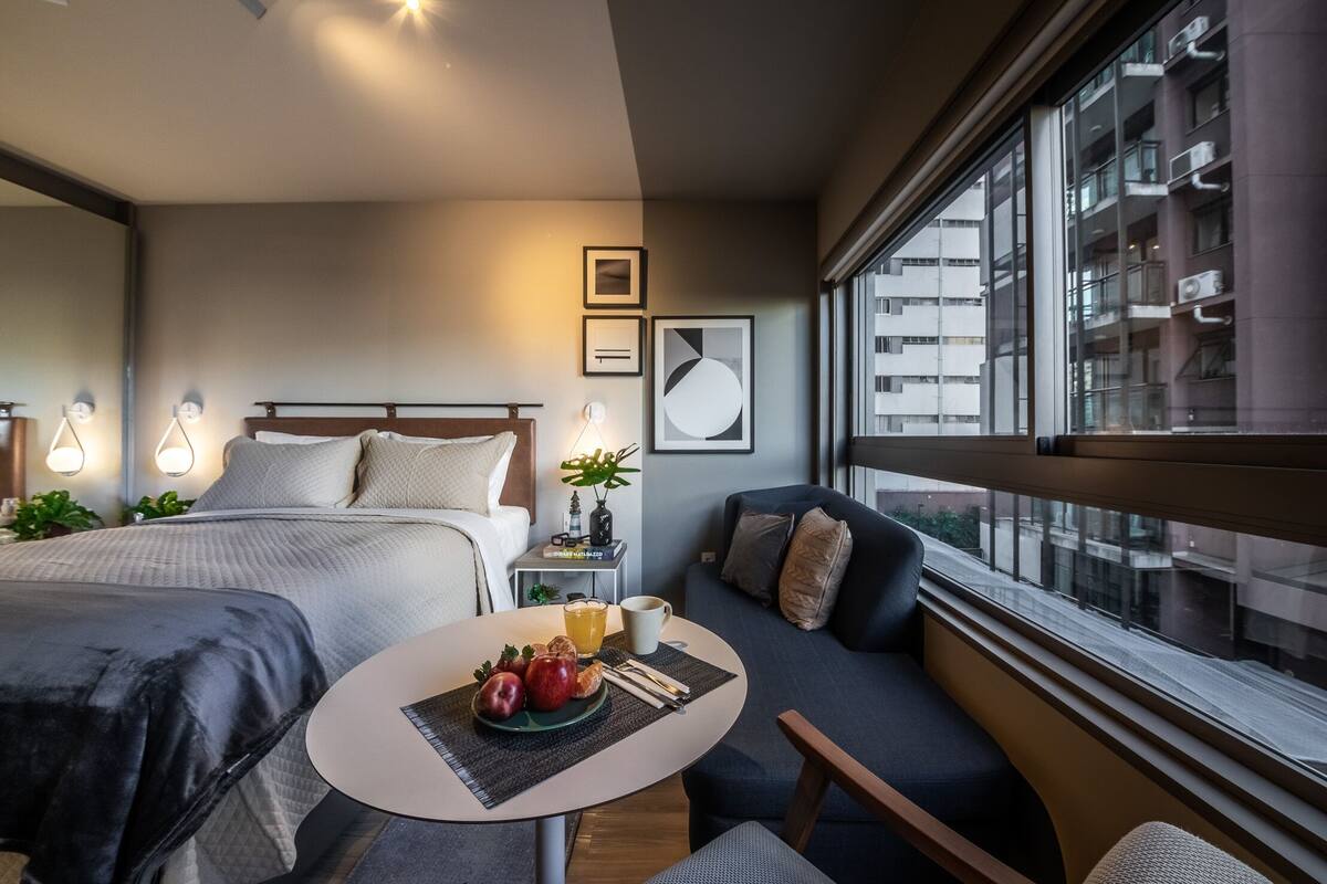 Una habitación de hotel moderna con una cama grande, un sofá, una mesa de comedor con frutas y una bebida, y vistas de la ciudad a través de amplios ventanales.