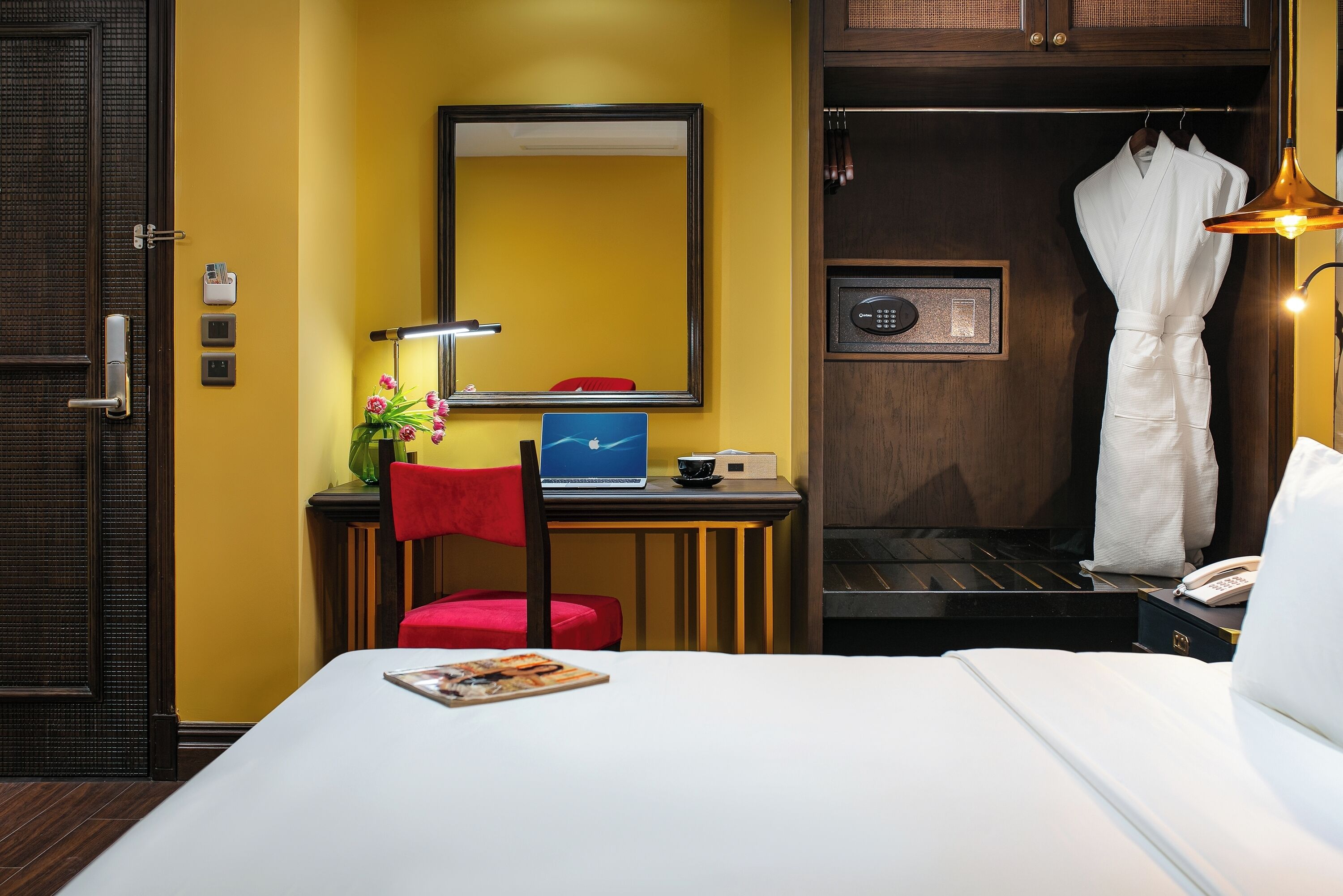 A hotel room with a bed, a desk with a laptop, a red chair, a mirror, a robe, and a phone.