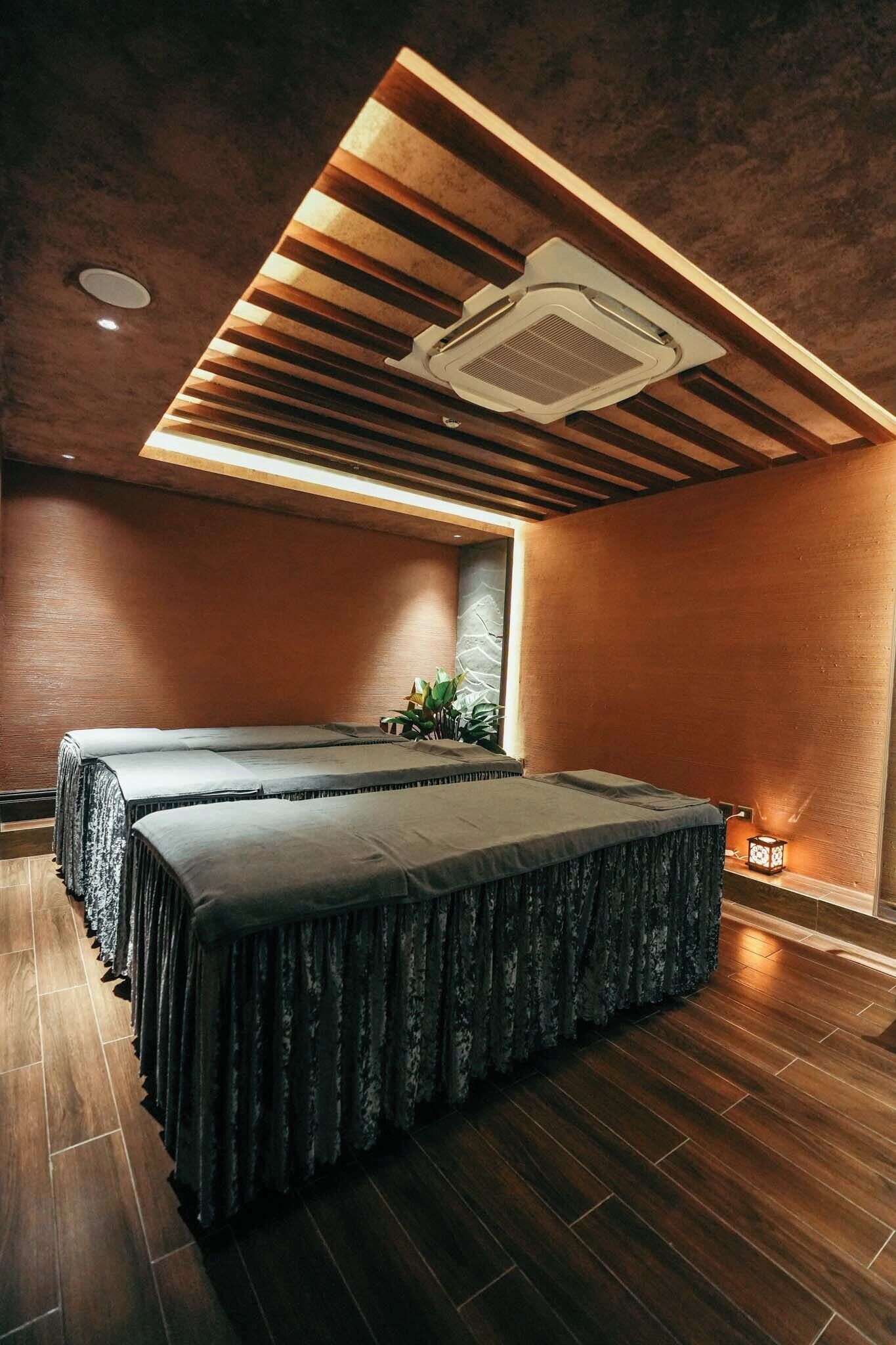A room with two beds, wooden flooring, a ceiling with a built-in air conditioner, and a small window with a plant.