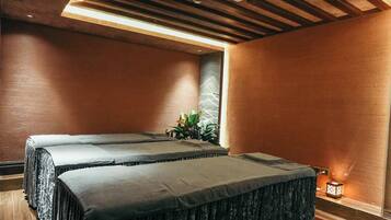 A room with two beds, wooden flooring, a ceiling with a built-in air conditioner, and a small window with a plant.