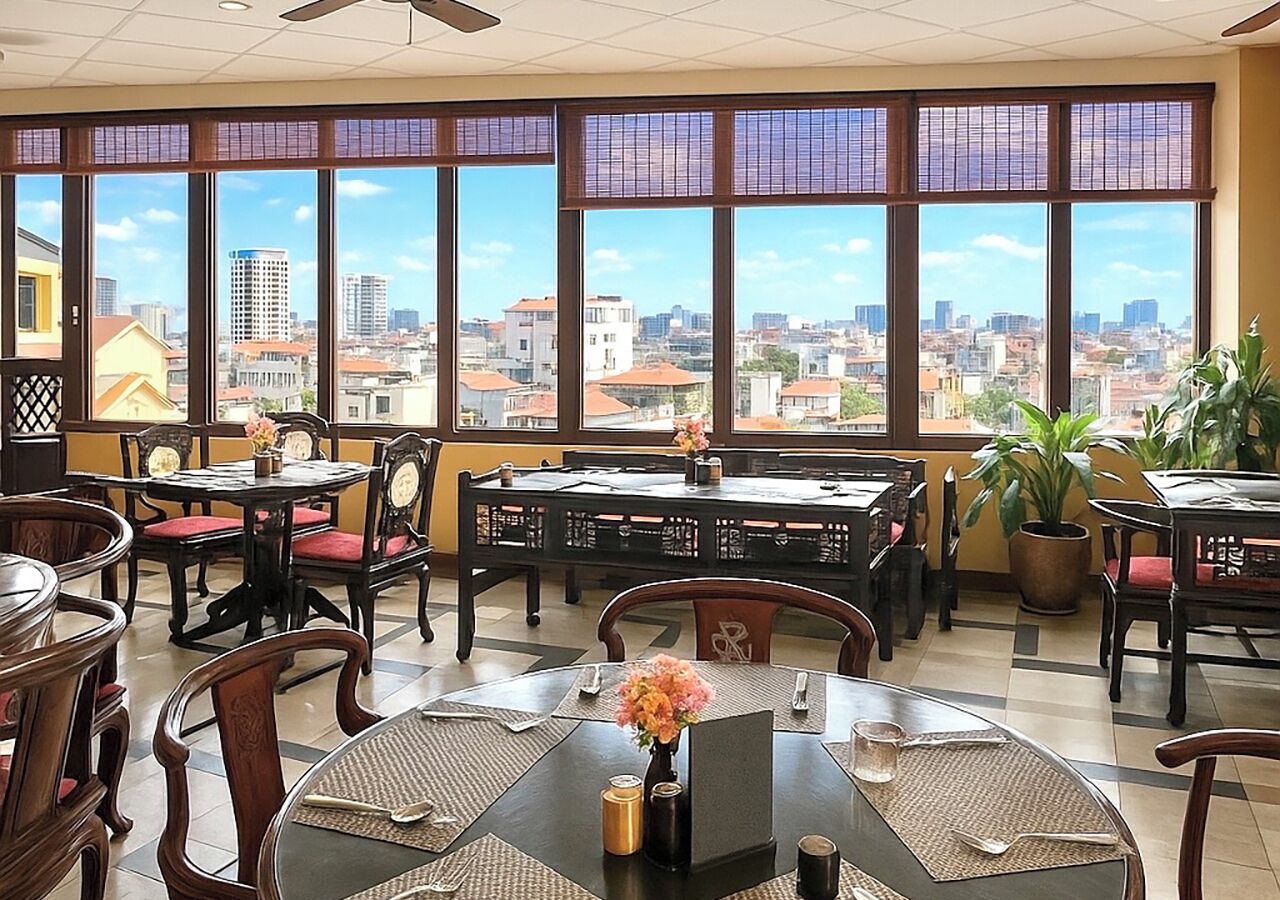 A restaurant with multiple tables and chairs, large windows with a city view, and potted plants.