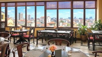 A restaurant with multiple tables and chairs, large windows with a city view, and potted plants.