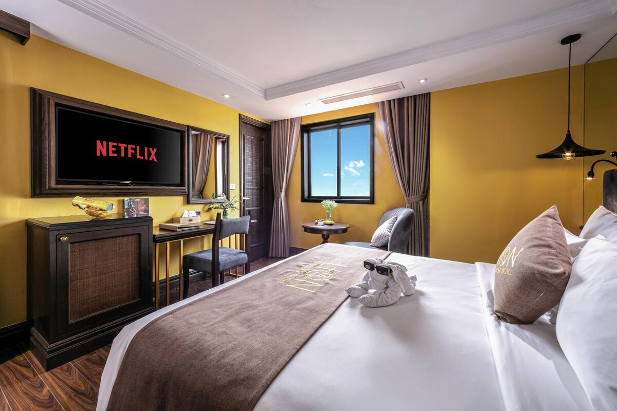 Habitación de hotel con una amplia televisión de pantalla plana, un escritorio, una silla, una cama con una manta y una ventana con cortinas.