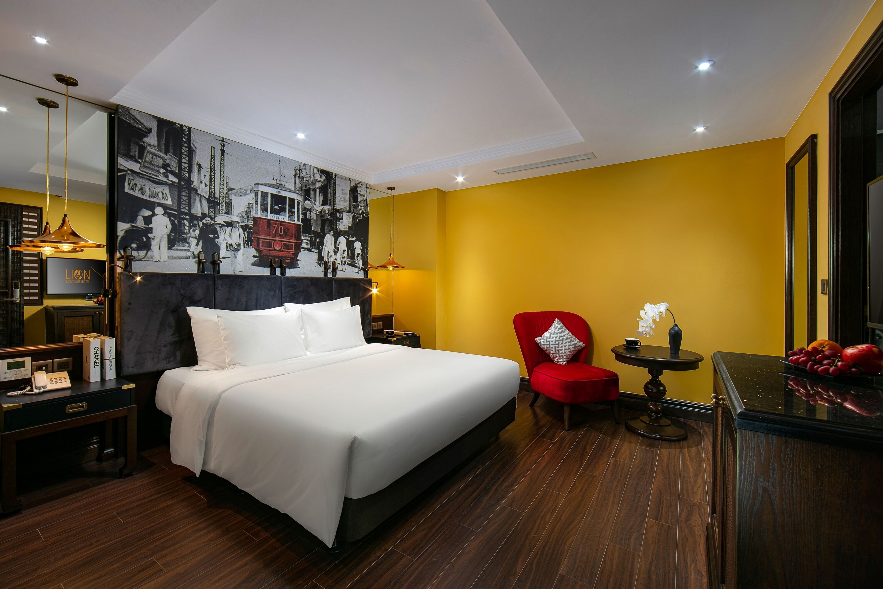 A hotel room with a large bed, a red armchair, a small table with a vase, and a wall-mounted artwork depicting a vintage tram.