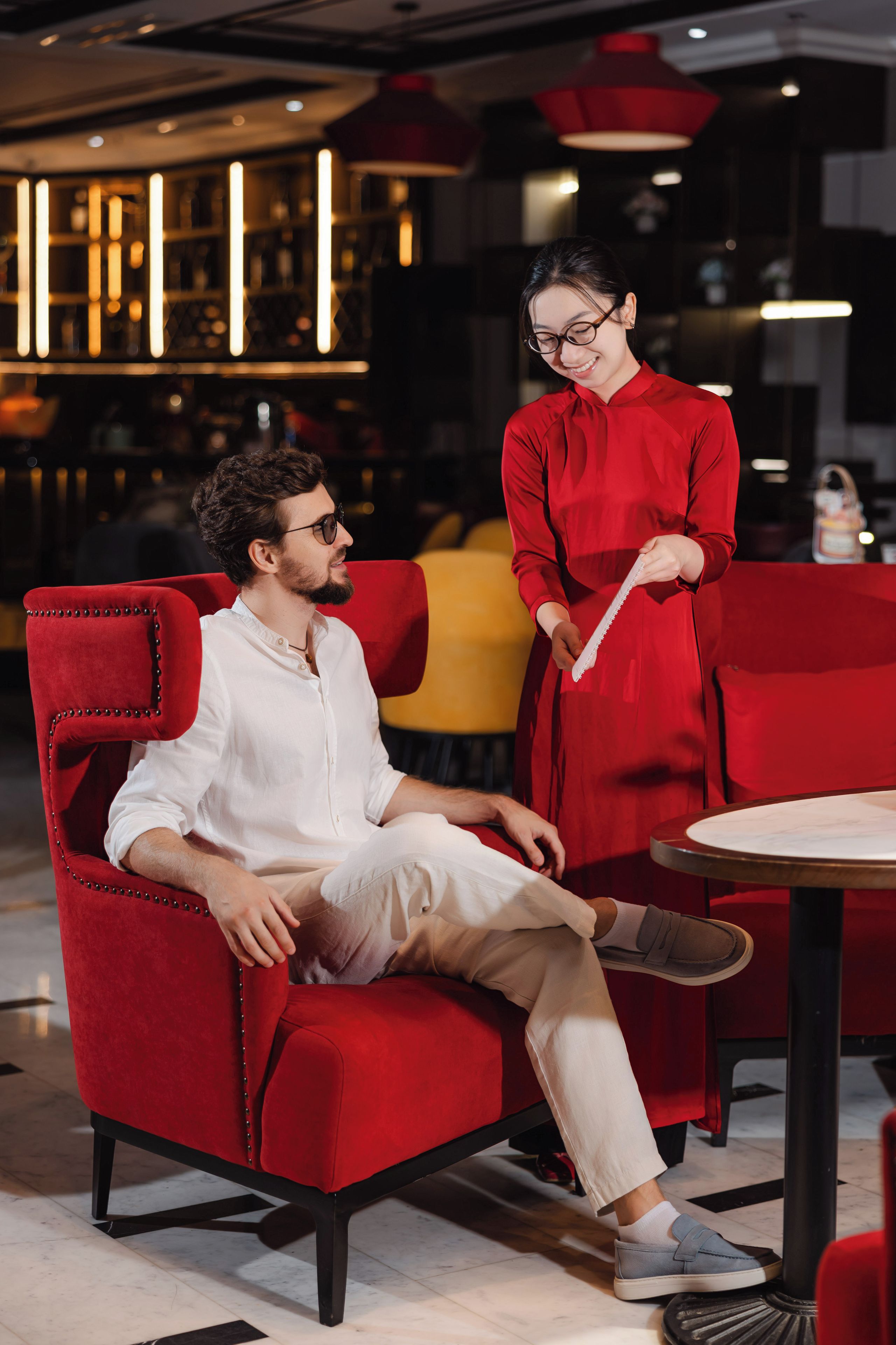 A man seated on a red upholstered chair in a modern interior with a woman standing beside him.