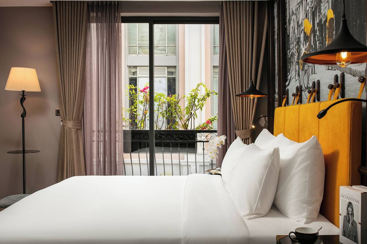 Habitación de hotel con una cama, una lámpara, una ventana con cortinas, vista al exterior y una pared con un cuadro y una estantería con libros.