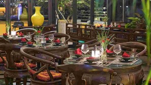 A restaurant with round tables set for dining, wooden chairs, and a large yellow vase centerpiece.