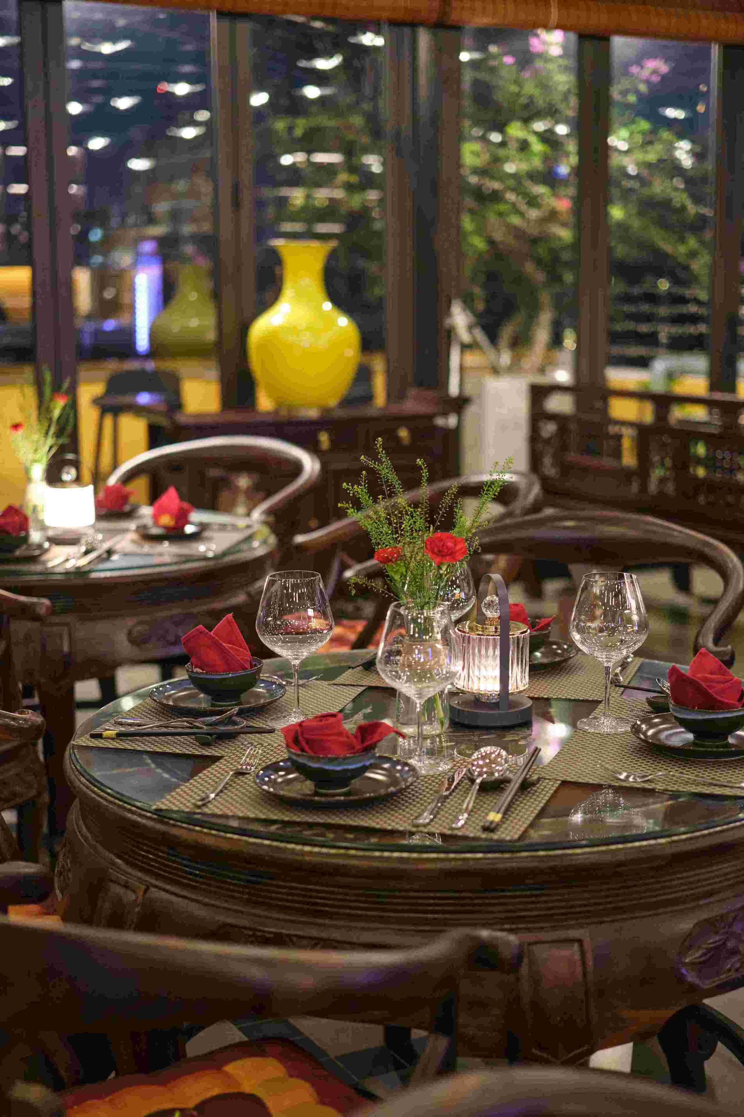 Un comedor con mesas redondas dispuestas para dos comensales, con copas, platos y servilletas rojas.