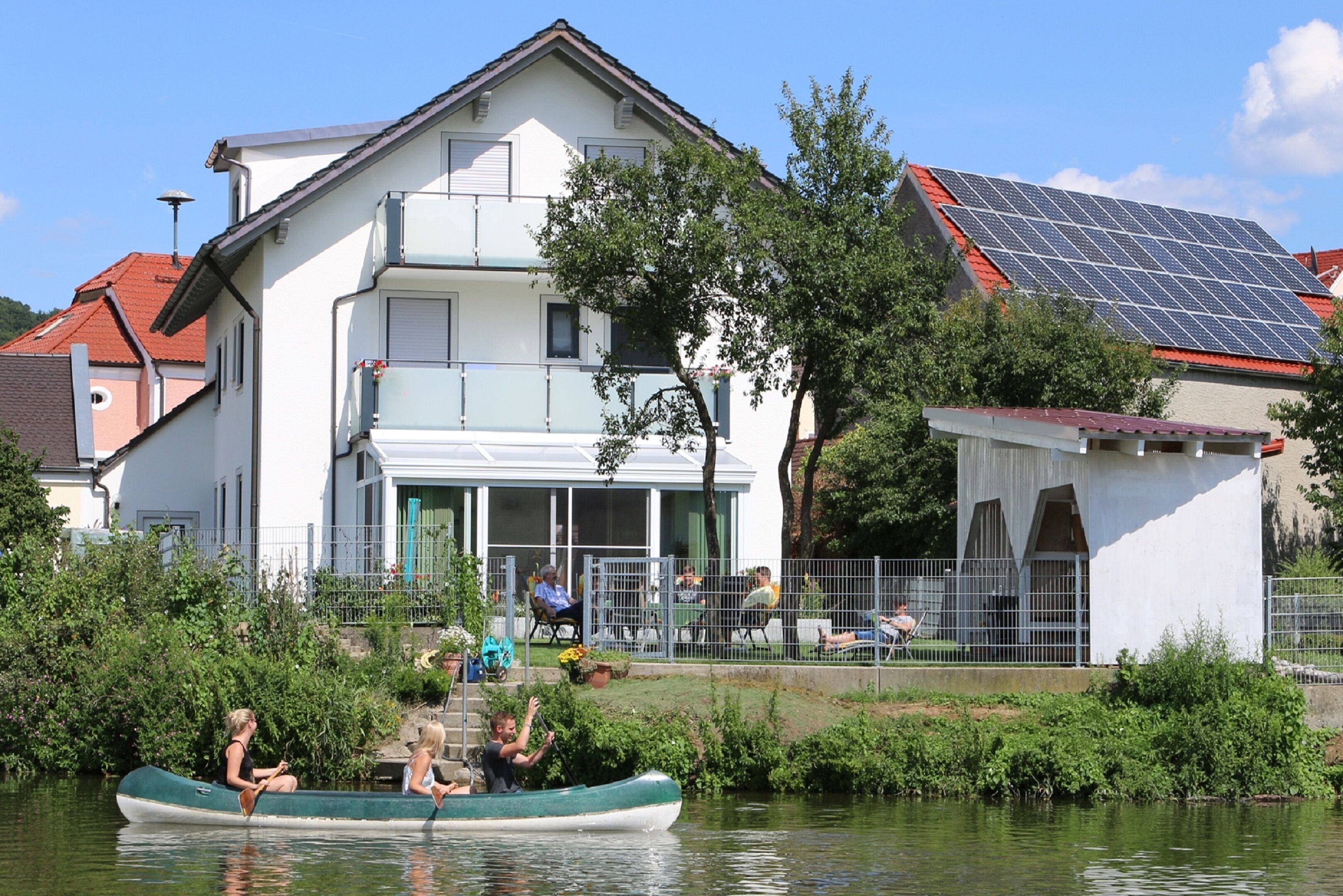 Una casa con balcón, panel solar y un río con un bote.