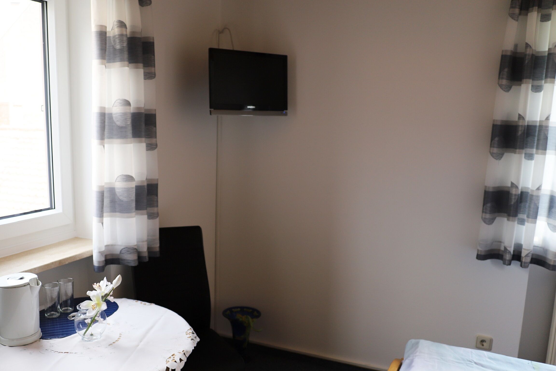 Ein Hotelzimmer mit Flachbildfernseher an der Wand, einem kleinen Tisch mit Blumenvase, einem Sessel und einem Bett mit blauer Matratze.