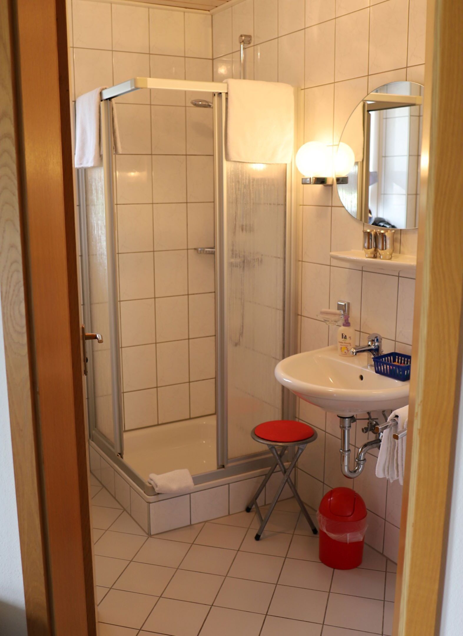 Un baño con cabina de ducha de vidrio, un lavabo con espejo y un taburete rojo.