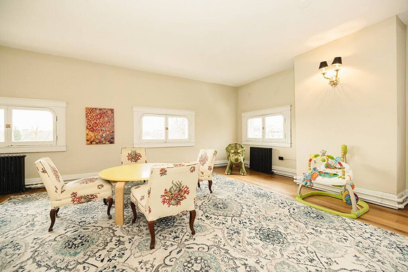 Une pièce spacieuse avec une table ronde et des chaises à motifs floraux, un tapis à dessins, un radiateur et un transat pour bébé.