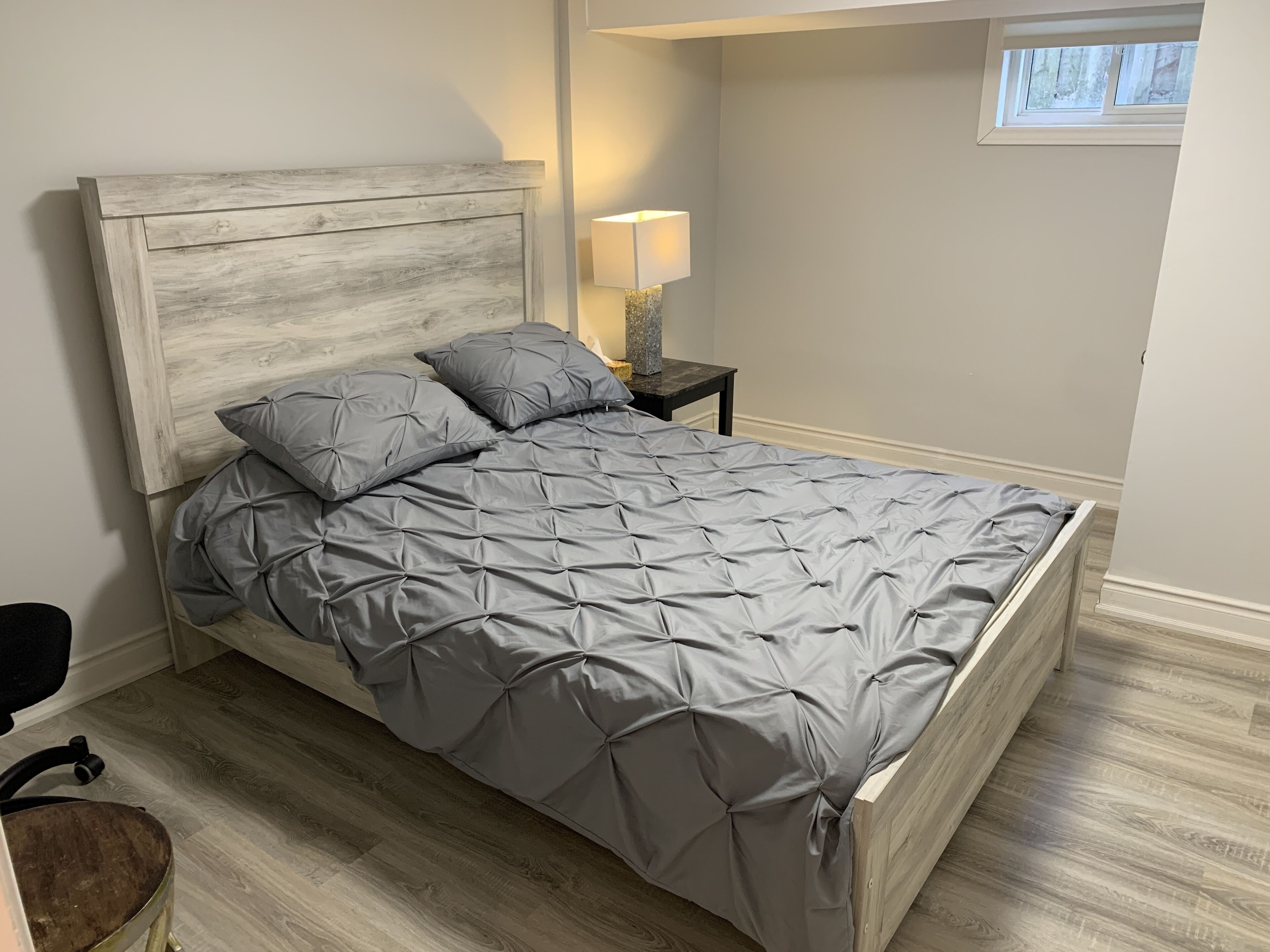 A bed with a wooden headboard, grey bedding, a nightstand with a lamp, and a window with a view of trees.