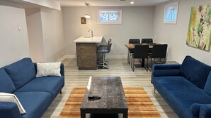 A living room with two blue sofas, a central coffee table, a dining area with a table and chairs, a kitchen area with a bar stool, and artwork on the walls. - Luxury 2 bedroom house - fully renovated - full feature (Richmond Hill)