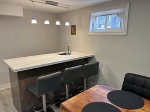 Une cuisine moderne avec un comptoir en marbre, trois tabourets de bar, une table à manger en bois avec des sets de table noirs et un éclairage suspendu.