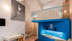 A modern living room with a blue accent wall, a dining area with a white table and chairs, and a kitchen area with blue cabinetry. - Architect designed studio (EP7L). Green rooftop! (Athina)