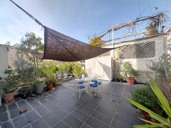 A patio area with a table and chairs, surrounded by potted plants and a shaded area.