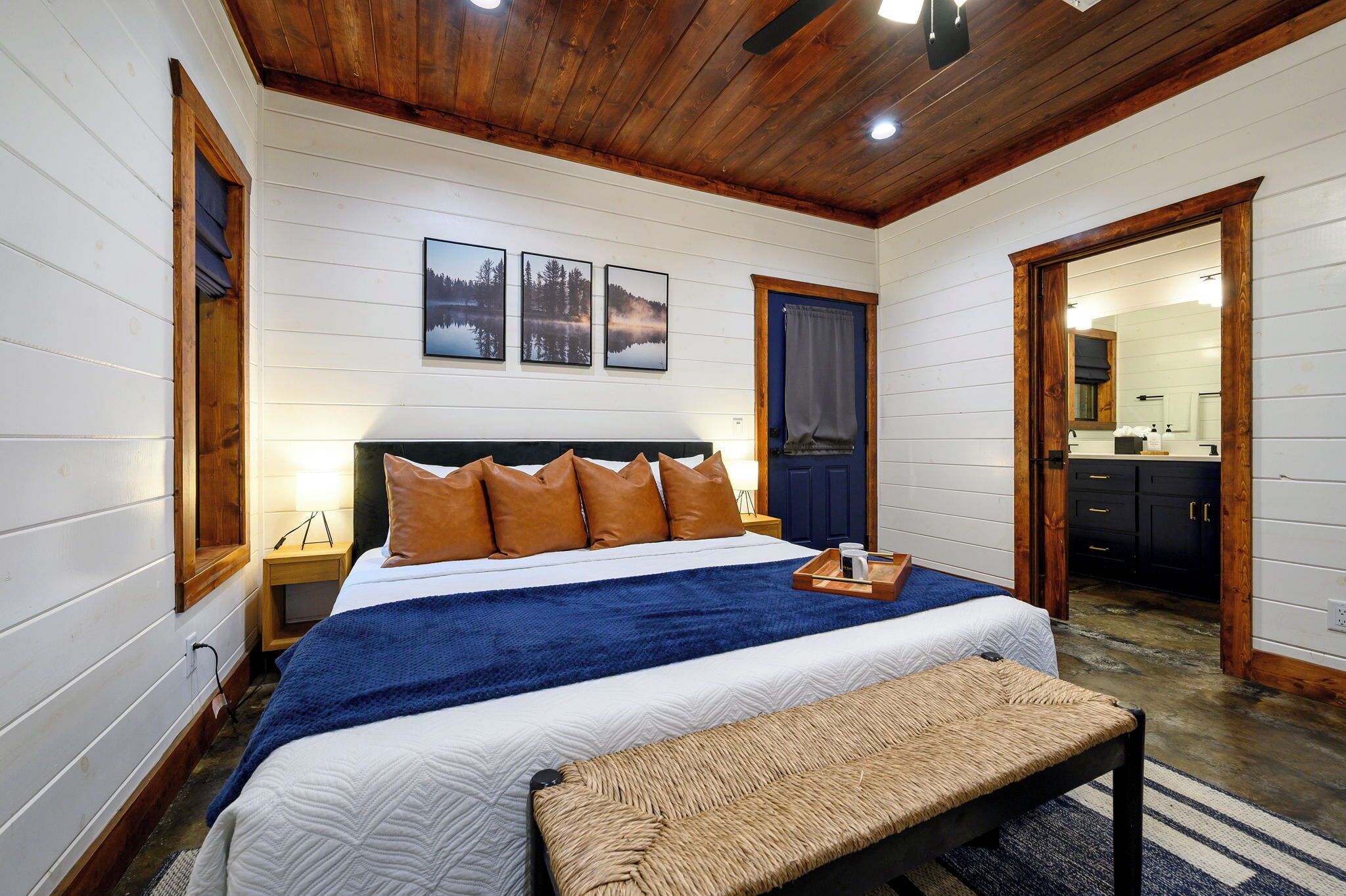 Une chambre avec un plafond en bois, un lit recouvert d’une couverture bleue, un banc en bois et une table de chevet avec une lampe.