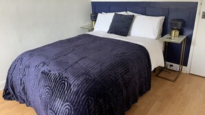 Cama bem arrumada com colcha azul escura estampada, duas almofadas e cabeceira combinando. Há duas mesas de cabeceira com abajures, e o quarto tem parede em tom claro com espelhos em formato de raios de sol.