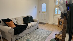 Uma sala de estar com um sofá bege, uma poltrona verde, um aparador de madeira e uma janela com persianas.