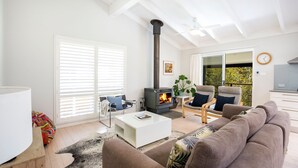 A modern living room with a fireplace, a sofa, a coffee table, and a large window with blinds. - Mariners Retreat - by Latitude South Coast (Vincentia)