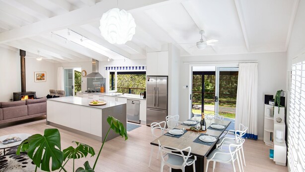 A modern kitchen with a central island, a dining area with a table set for four, and a living area with a sofa and a fireplace. - Mariners Retreat - by Latitude South Coast (Vincentia)