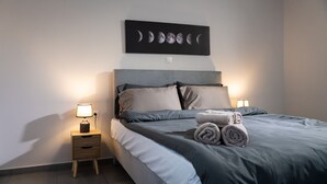 A neatly made bed with grey bedding and two pillows, a bedside table with a lamp, and a wall-mounted artwork depicting moon phases. - Andromachis Moneto Under Meteora (Kalabaka)