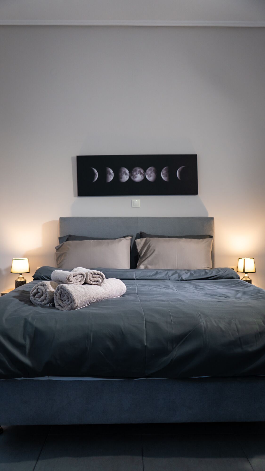 Una cama bien hecha con toallas enrolladas, un calendario de fases lunares en la pared y dos lámparas de noche.
