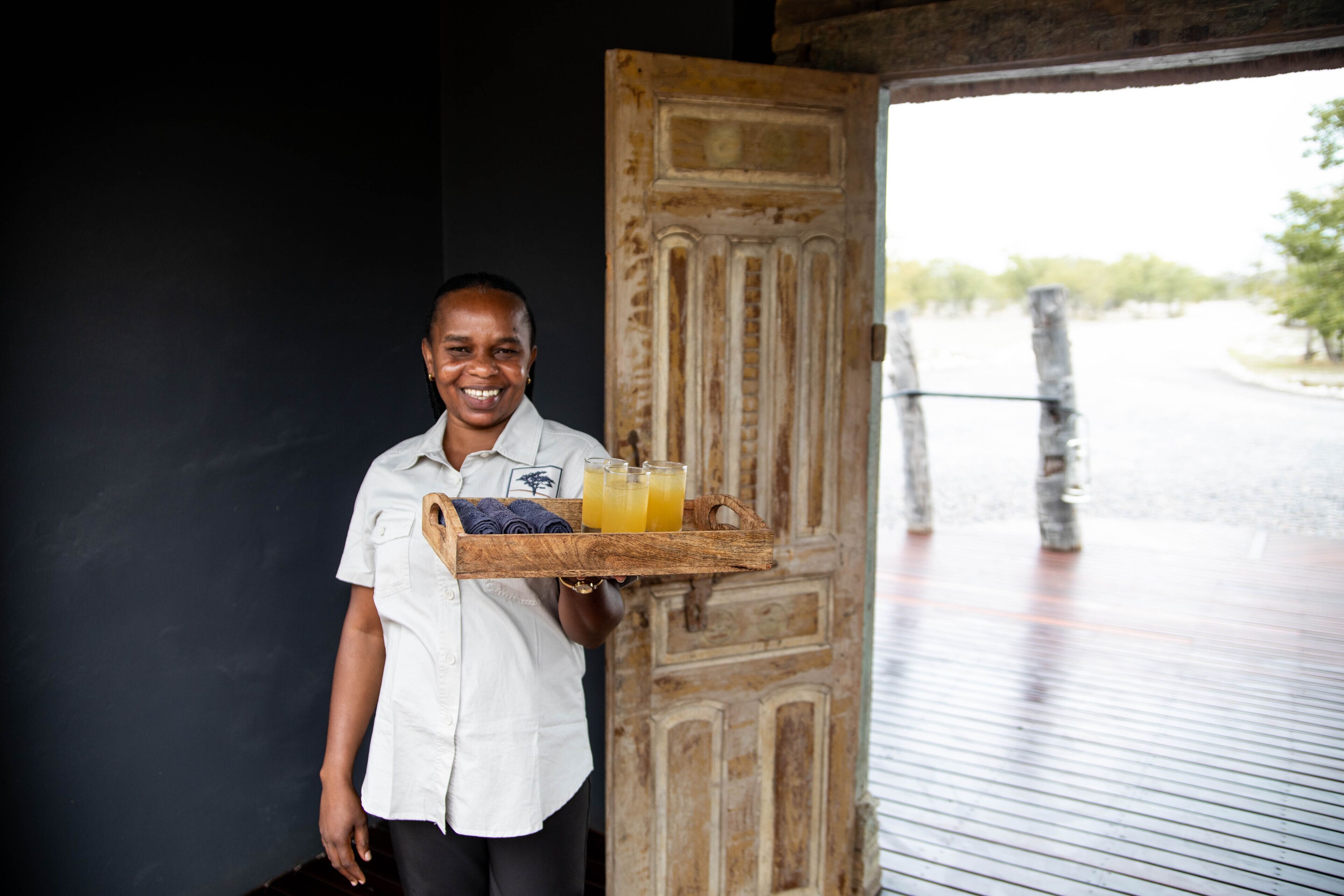 A person holding a tray with drinks and a towel, standing in front of a wooden door with a view of a water body and trees.