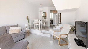 Una sala de estar moderna con un sofá gris, un sillón blanco y un televisor. Se ve una escalera que lleva a una zona de cocina.
