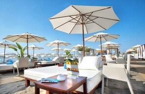 Un espace de détente aménagé sur la plage, avec plusieurs chaises longues blanches et de grands parasols, une table en bois sur laquelle est posé un menu, et une vue sur la mer.