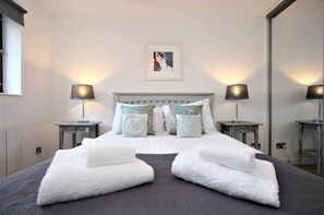 Una cama bien hecha con toallas blancas, cabecera gris y dos lámparas de noche.