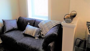 A dark grey sofa with a striped throw blanket and two pillows, positioned next to a window with a view of a building. A metal basket is placed on a white cabinet to the right. - Newly Renovated "Arctic Rose" Loft in Historic Florence (Florence)