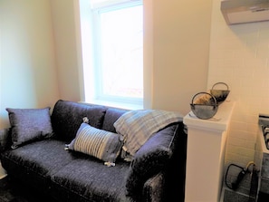 A dark grey sofa with a striped throw blanket and two pillows, positioned next to a window with a view of a building. A metal basket is placed on a white cabinet to the right.