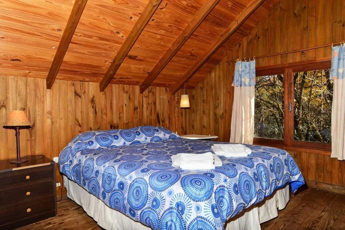A wooden cabin bedroom with a bed, a nightstand, a lamp, and a window with curtains.