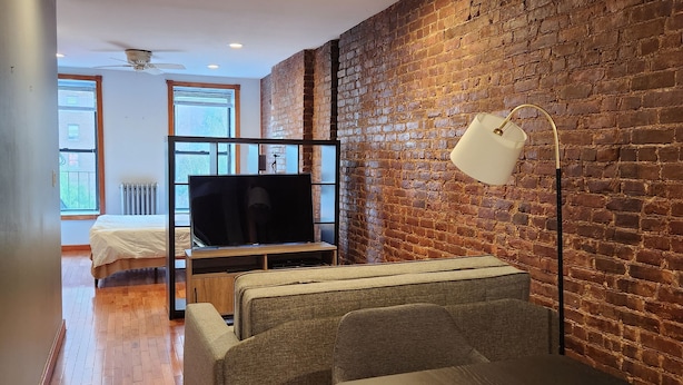 A modern living room with a brick wall, wooden flooring, a ceiling fan, and a television. - Studio boutique à 2 min do Central Park (New York)