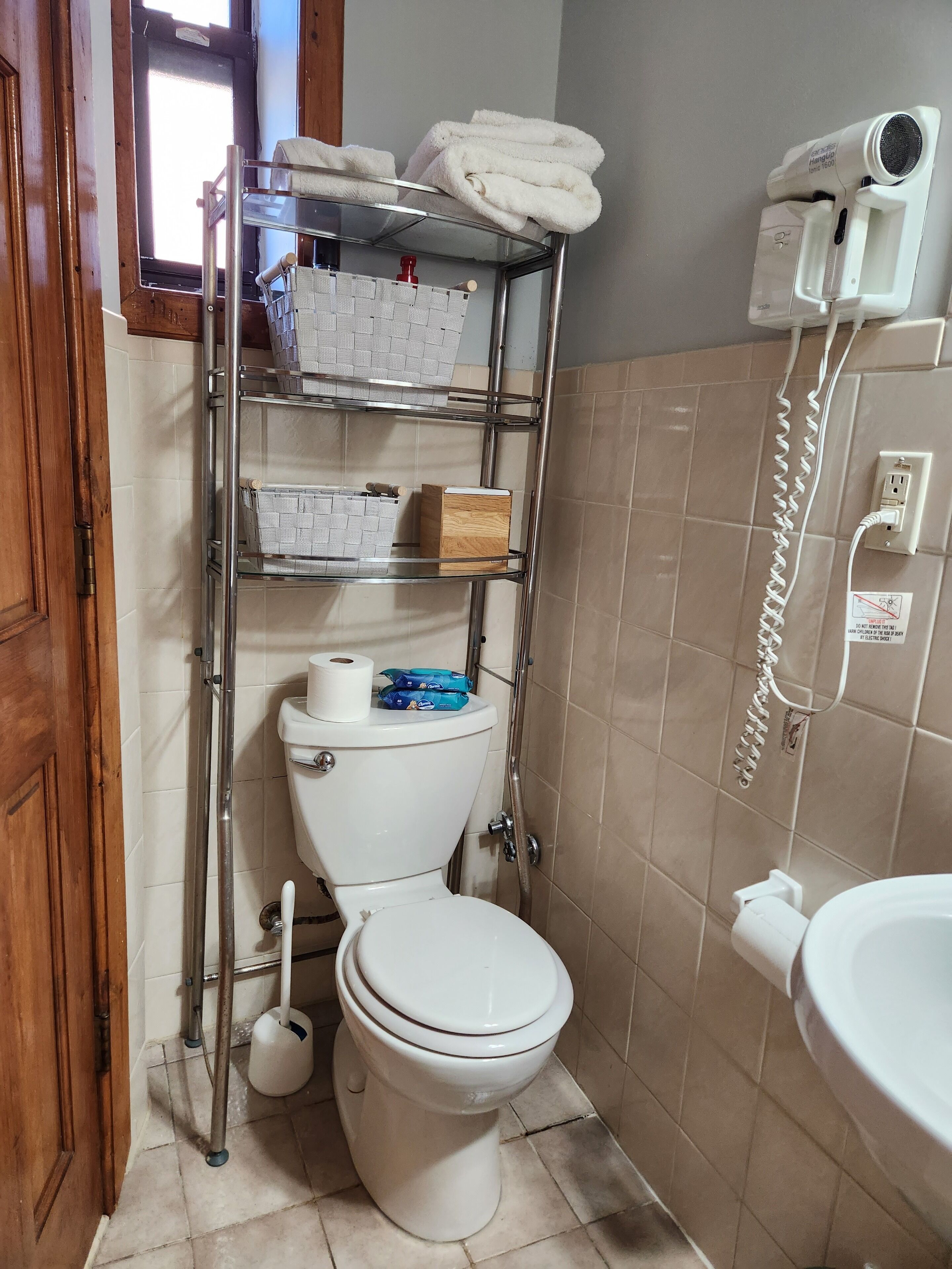 A bathroom with a toilet, a shelf with towels and toiletries, a hanging toilet paper holder, and a phone on the wall.