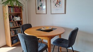 Ein Essbereich mit einem runden Holztisch, zwei grau gepolsterten Stühlen, einem Bücherregal mit Büchern und einem Radio, sowie zwei gerahmten Kunstwerken an der Wand.